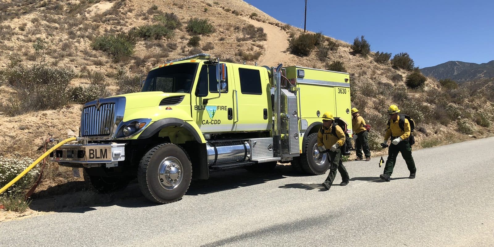 Three firefighters in yellow shirts and helmets pull hose from a lime-green wildland fire engine parked on a road below a dry, brush-covered hillside.