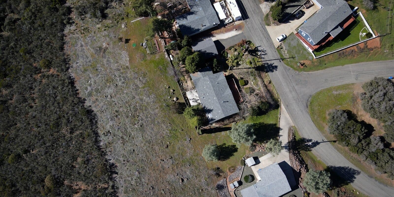 An aerial view looking straight down at homes whose back yards meet a strip of dry grass and dense chaparral.