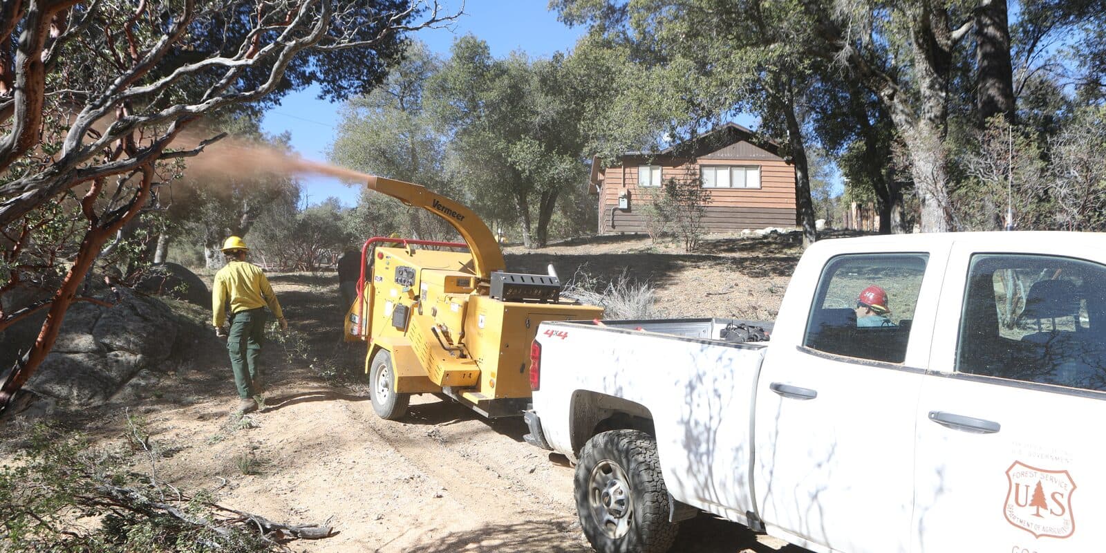A fuels crew member walks beside a wood chipper next to a Forest Service truck, with a cabin among the oaks behind them.