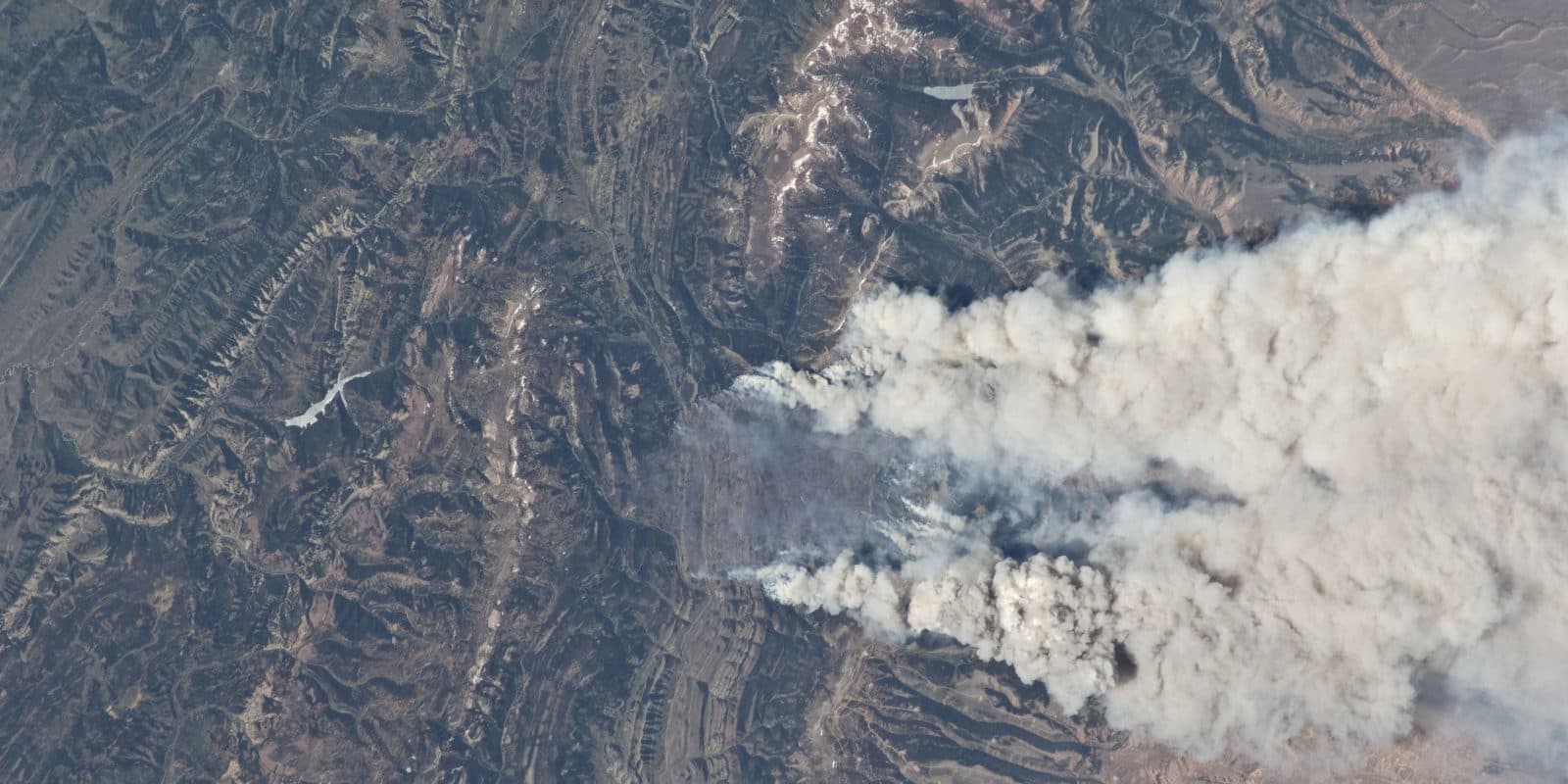 An overhead view from orbit of smoke plumes drifting across ridged terrain.