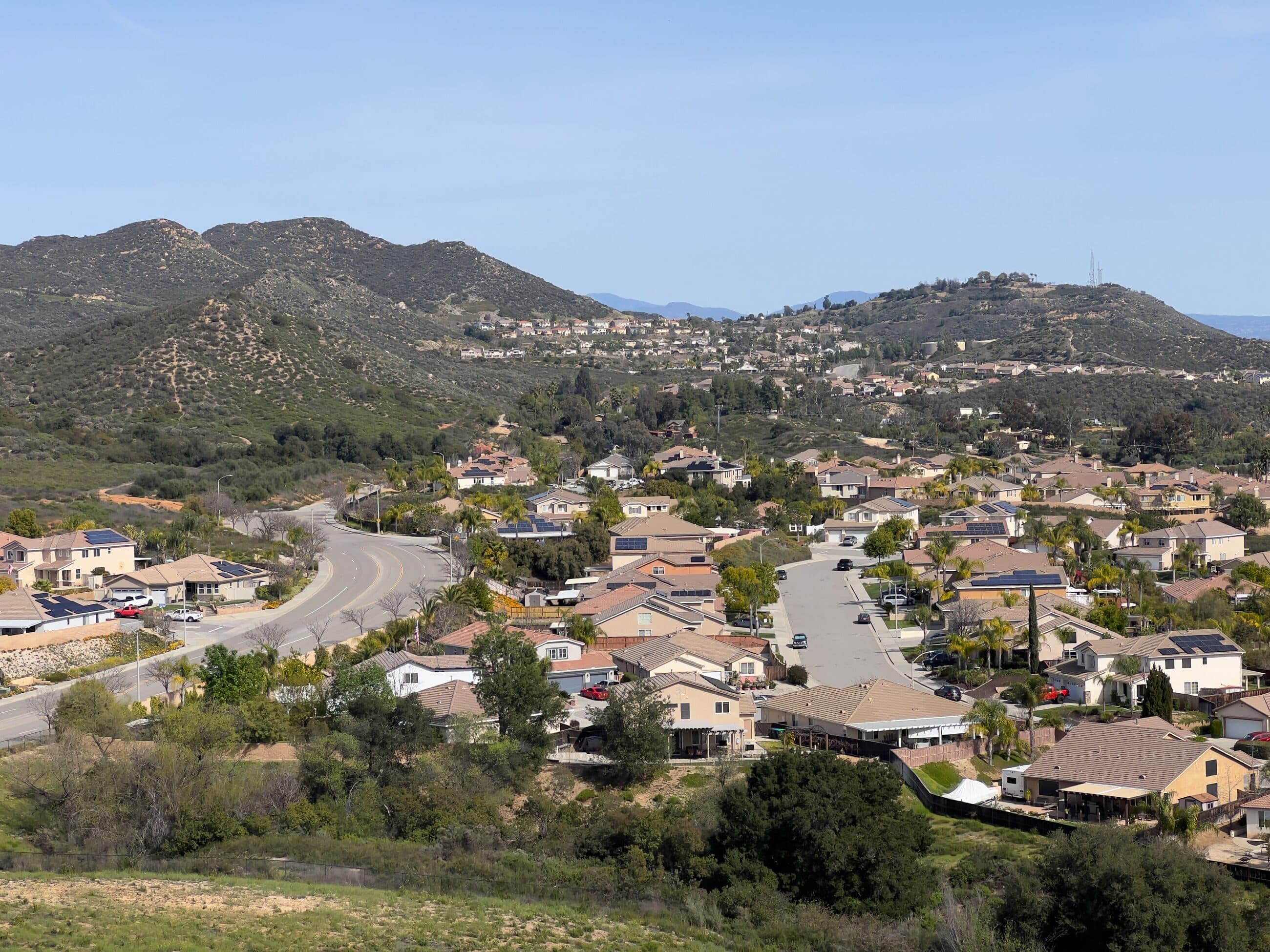 A neighborhood of homes fills a valley below chaparral-covered hills, with more houses climbing the slopes behind.