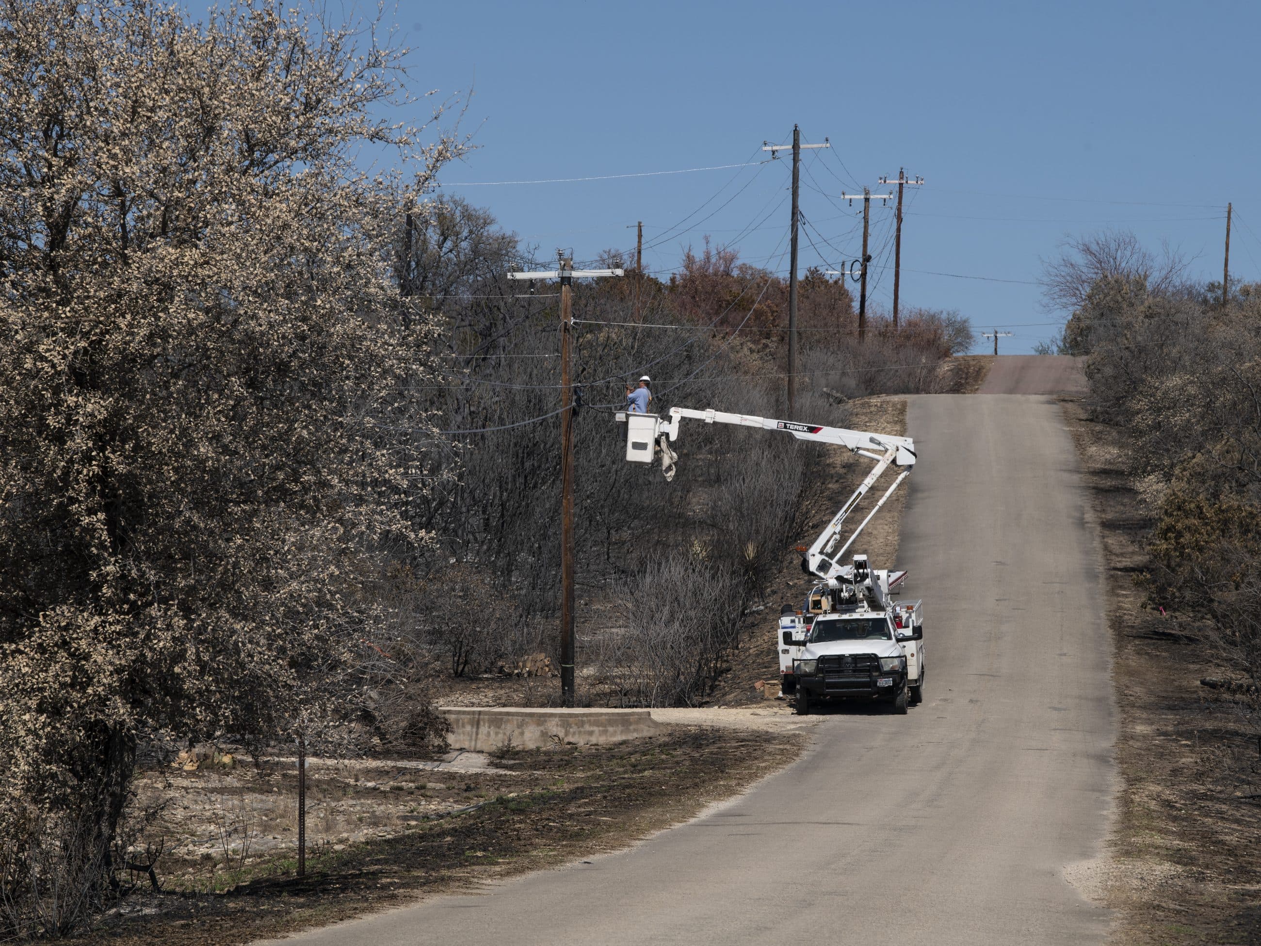 A utility line crew working from a bucket truck to restore power lines along a road running through burned ground.